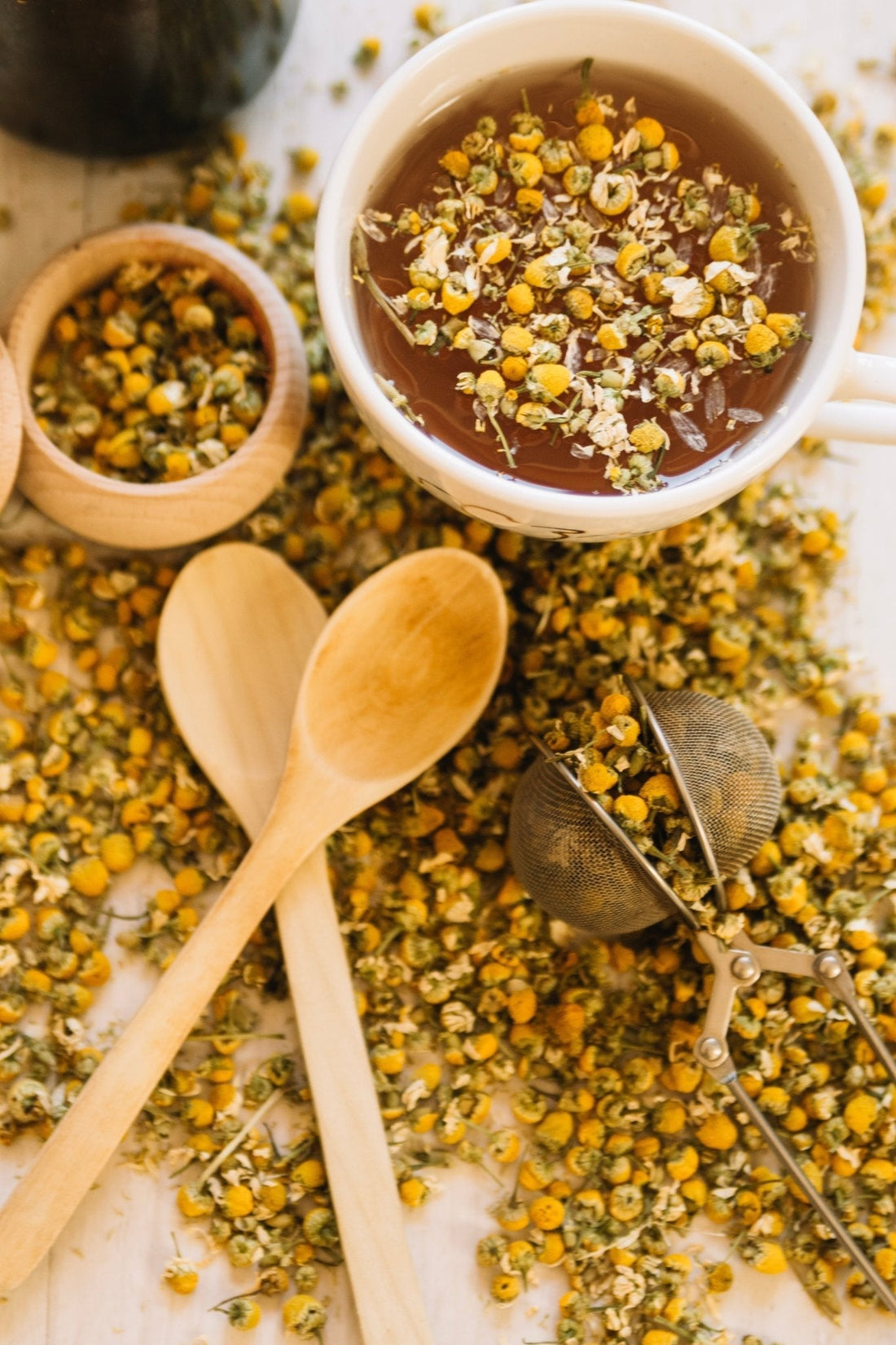 Chamomile tea cup surrounded by dried chamomile flowers and wooden spoons showing chamomile benefits for skincare