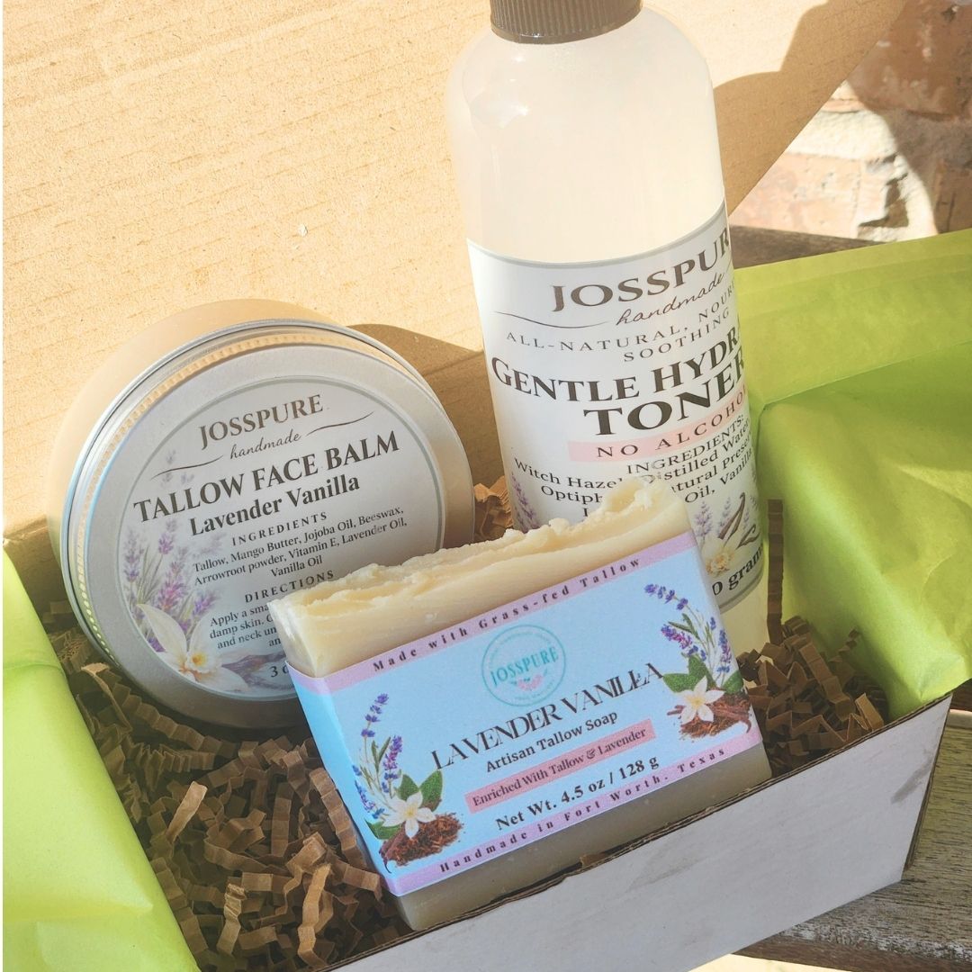 A Josspure gift set containing a lavender face balm in a tin, a gentle hydrating toner in a bottle, and a balance body wash bar, arranged in a wooden box with brown paper shred and green tissue paper.