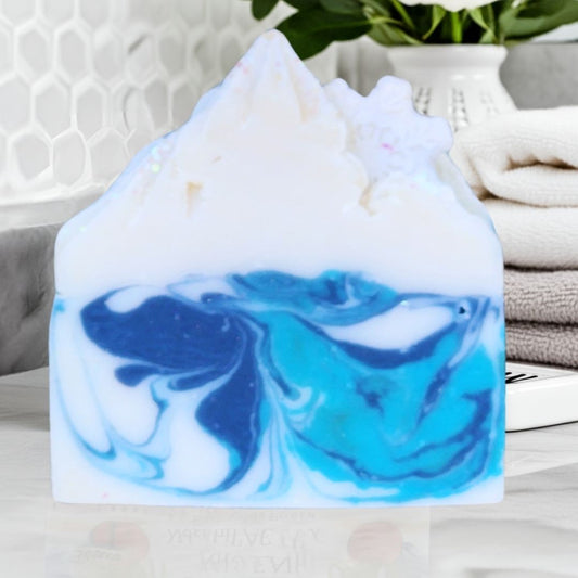 A handmade mountain-shaped soap bar with a white top and swirling blue and teal marbled swirls on the bottom, displayed on a light surface with a blurred bathroom background.