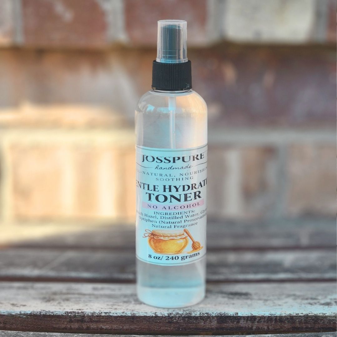 A bottle of Josspure Handmade Gentle Hydrator Toner with a black spray cap, placed on a weathered wooden surface with a blurred brick wall background.