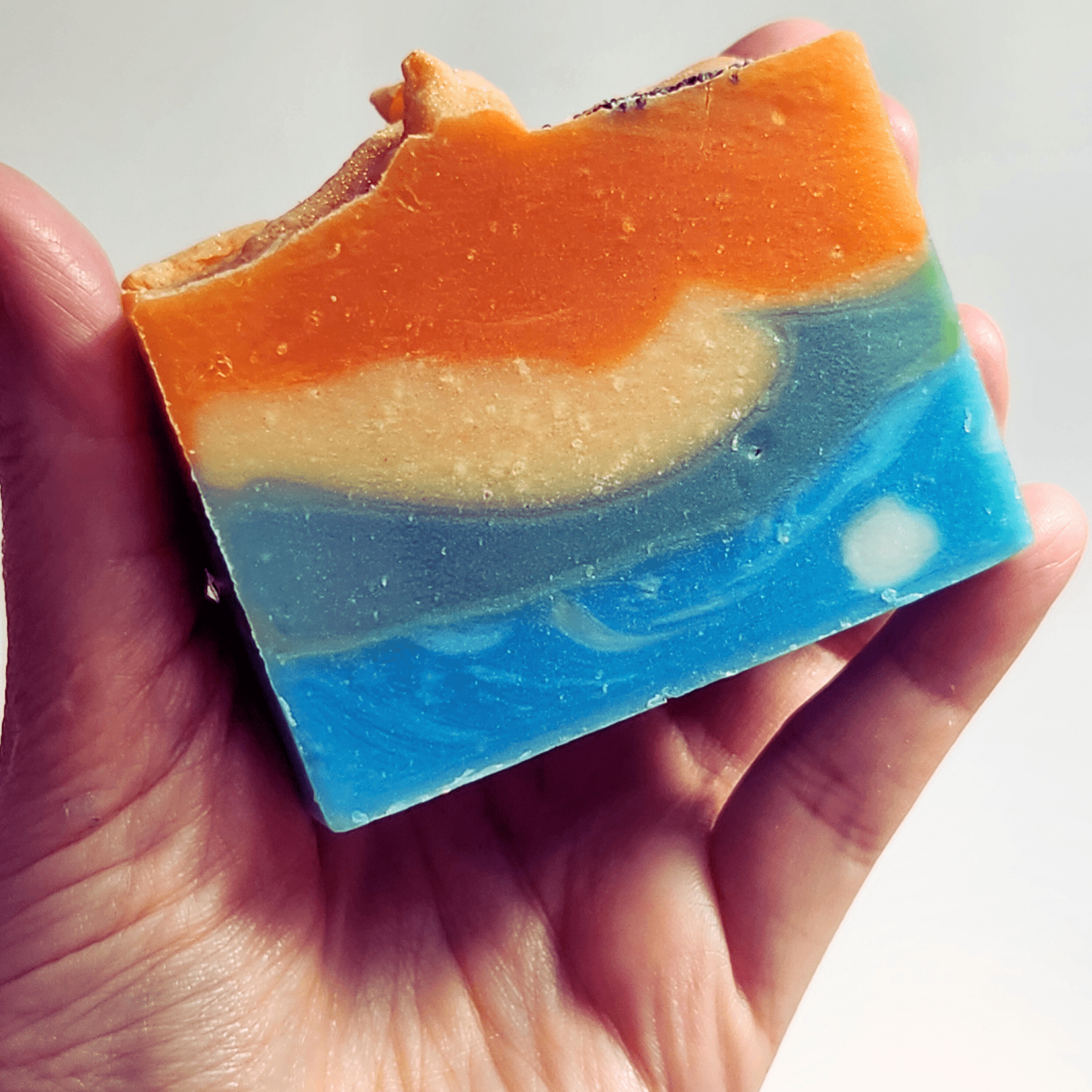 Hand holding a colorful mango papaya shea butter soap with orange, cream, and blue layers