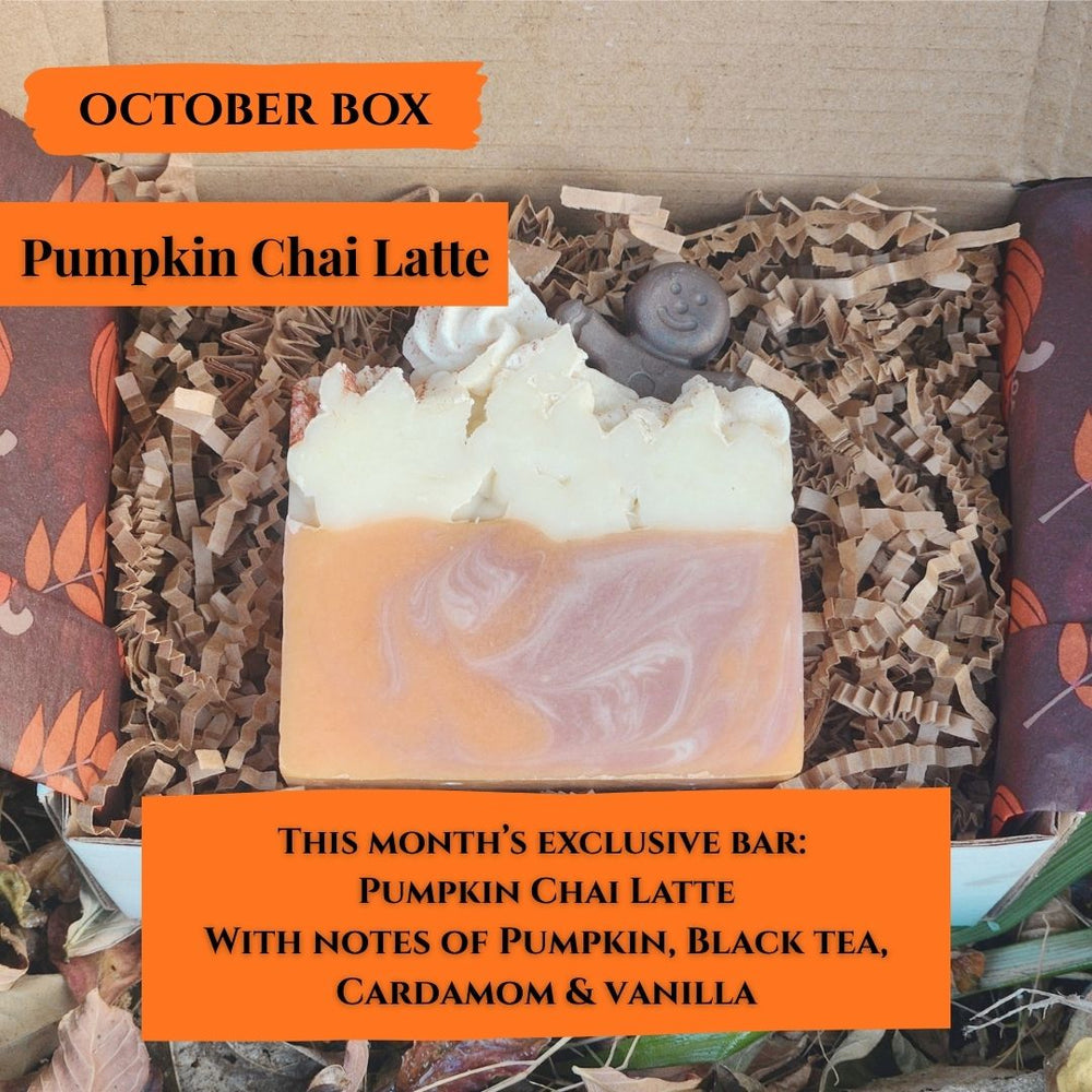 A bar of Pumpkin Chai Latte soap displayed inside an open cardboard gift box filled with brown crinkle paper. The soap has a marbled design with a light orange lower section and a white textured top resembling foam.