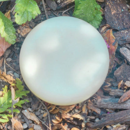 A round, frosted glass orb with a pale mint green tint, resting on a bed of mulch and green leaves outdoors.